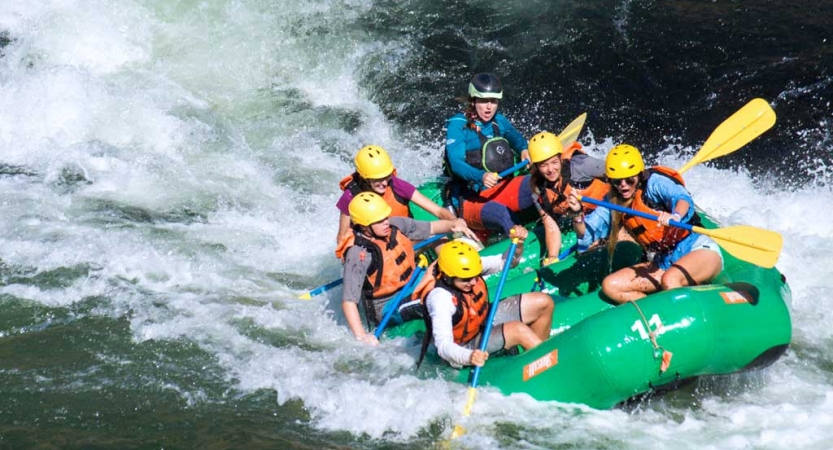 a group of students wearing helmets and life jackets paddle a green raft through whitewater